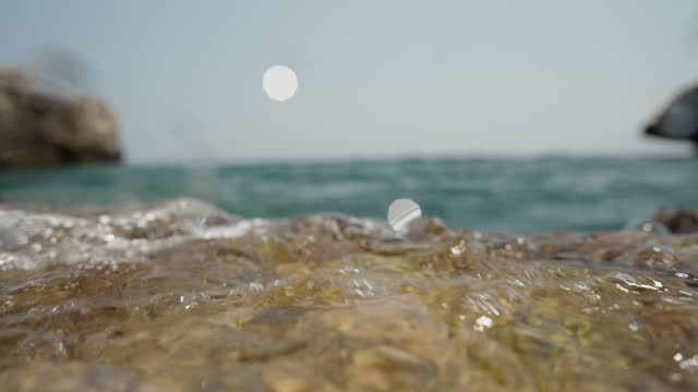 The camera lies on a rock at the edge of the sea, and a wave washes over it, in slow motion, capturing a close-up view.