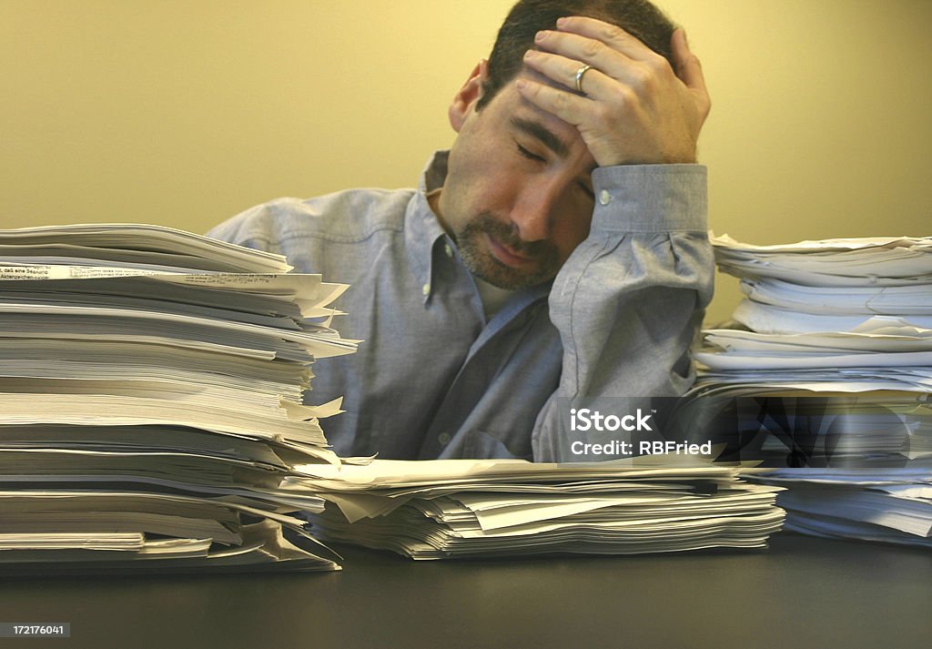 Headache a frustrated man holding his head in the background, with stacks of papers in the foreground (focus on the stacks of papers) Adult Stock Photo