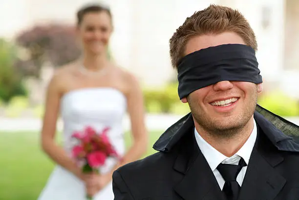 Humorous wedding photograph of a lovely couple. The groom has been blindfolded to ensure he cannot see his beautiful bride before the wedding. The bride stands in the background making sure it is safe before she walks by.