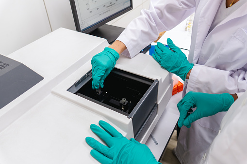 Professional technician putting sample container containing liquid solution into specimen holder of laboratory equipment for analysis while colleague standing together for observation