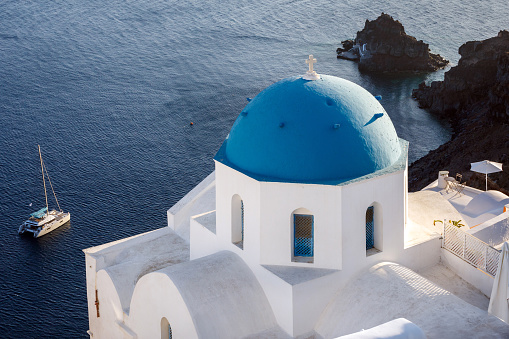 A gorgeous view of a quant church and blue sea with sailing boat on Oia, Santorini, Greece.