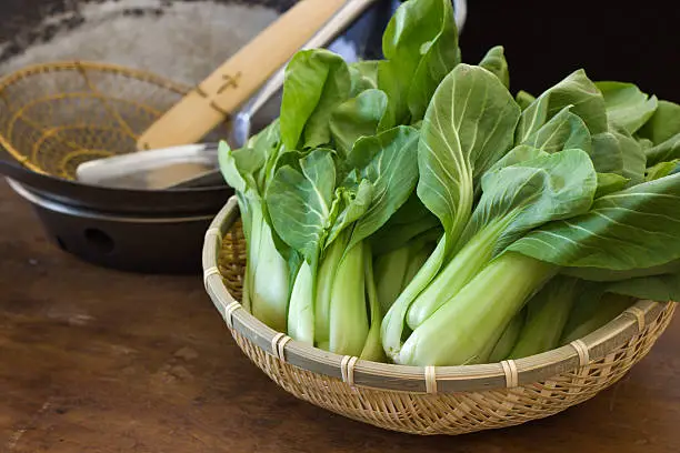"Subject: A basket of baby bok choy, a common Chinese vegetable, and a Chinese wok, resting on a kichen counter in preparation for cooking."