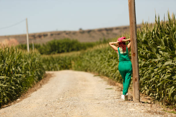 Woman with hat walking along dirt road between planted corn field, on her private farm
