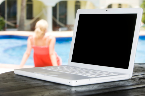 Laptop close up on outdoor table as young blond woman relaxes in background by pool. CLICK FOR SIMILAR IMAGES AND LIGHTBOX WITH MORE PEOPLE USING LAPTOPS.