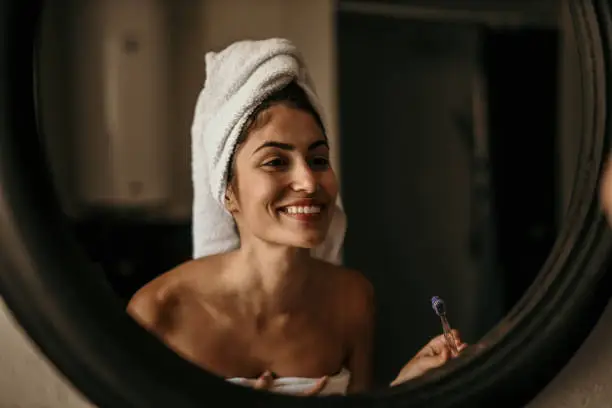 Photo of Latin beauty embraces her morning routine, wrapped in a towel and brushing her teeth in front of the bathroom mirror, radiating confidence and self-assuredness.