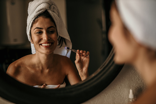 Latin beauty embraces her morning routine, wrapped in a towel and brushing her teeth in front of the bathroom mirror, radiating confidence and self-assuredness.