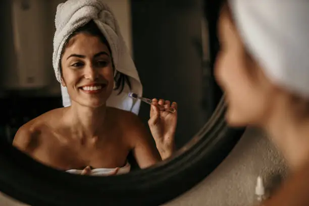 Photo of Latin beauty embraces her morning routine, wrapped in a towel and brushing her teeth in front of the bathroom mirror, radiating confidence and self-assuredness.