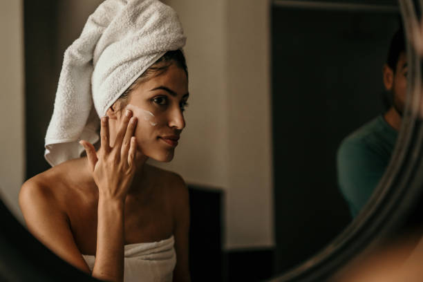 A young woman applies moisturizer to her face, her reflection in the mirror capturing her dedication to self-care stock photo