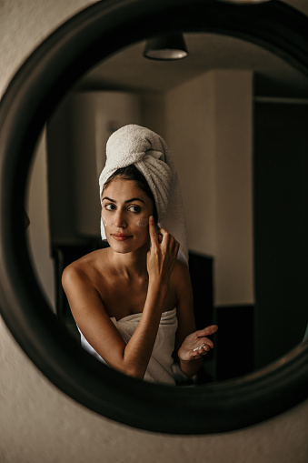 A young woman applies moisturizer to her face, her reflection in the mirror capturing her dedication to self-care