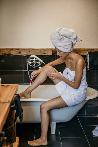 A woman sits in a bathtub, immersed in the act of shaving her legs, showcasing a moment of personal grooming
