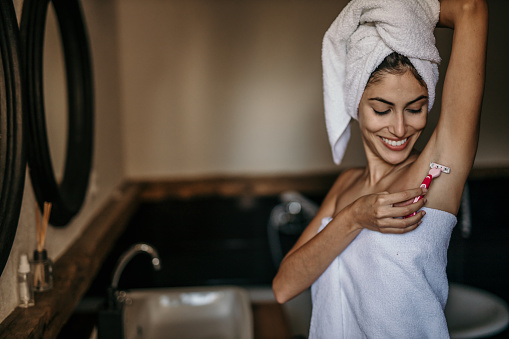 An elegant woman with a towel wrapped around her waist, meticulously shaving her underarms in her bathroom