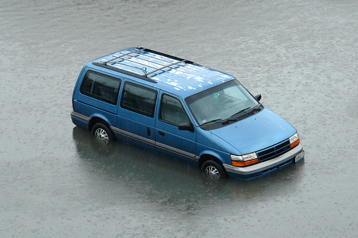   An abandoned van caught in the rising flood waters. No driver is visible through the windshield. The paint finish is peeling off the roof in some parts, but the vehicle is otherwise in good condition. The entire surroundings are consumed by the flood waters as a driving rain pelts the surface.