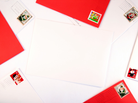 This color photo shows various red and white envelopes from the mail with Christmas postage, forming a holiday border around a large, central, blank envelope. The central envelope is white and awaits your text or design. The image background is white, and the image orientation is landscape.