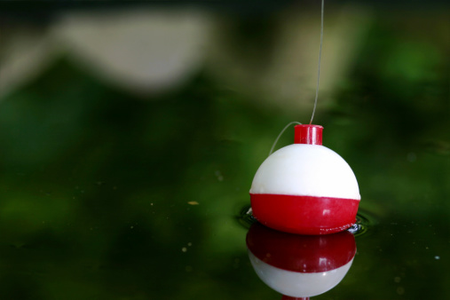Photo of a fishing cork in a pond.  Focus is on the top of the bobber were the fishing line is attached.  Bobber is also reflected in the water.