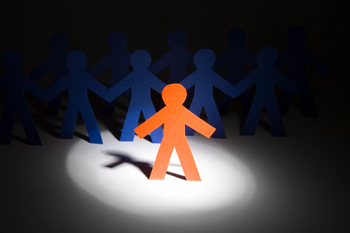 In the limelight: Orange paper person in a cone of light, with two rows of blue people in the shadows behind. Second row of people barely visible. Focus on the head and shoulders of the orange person. 