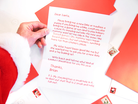 This mostly red color photo shows Santa’s red sleeve and hand holding a Christmas letter from a child named Brian at legible distance. The table or desk beneath the letter is covered with envelopes with Christmas postage, representing some of the thousands of letters he receives before the holiday each year.