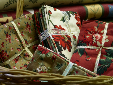 A collection of similarly toned fat quarter fabric bundles in a wicker basket; piled fabric bolts in background.