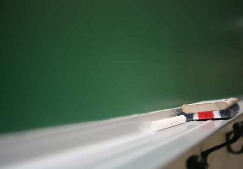 old technology - chalk and chalk eraser sit on steel ledge against green chalkboard in a school classroom. The brush is red, white and blue, with a wooden handle. The chalk is white. A couple of coat hooks are visible beneath the ledge.