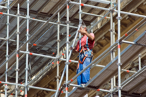 Construction worker who has dark skin and grey hair is up on scaffolding working on the exterior of an old building in the daylight. The construction worker is wearing a red t-shirt and blue working pants dealing with iron pipes of the building.