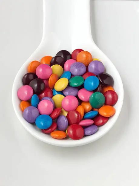 A white spoon full of bright candy coated chocolate sweets.
