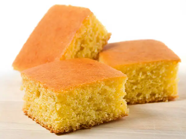 Three crumbly delicious homemade squares of melt in your mouth cornbread sitting on top of a birch cutting board.  These pieces are just begging for some sweet cream butter and clover honey to smother them.