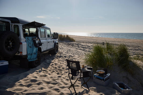 Land rover Defender 4x4 camping on the beach Domburg. It is a beautiful summer day by the sea, camp is outside in the sand and there is a small stove where a meal is prepared. The car stands between the grass in the sand.