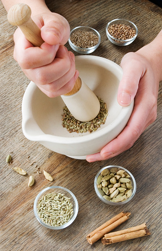 Grinding spices in a pestle and mortar on a wooden table top. The spices visible are Cardomom,fennel,cinnamon,coriander seeds.