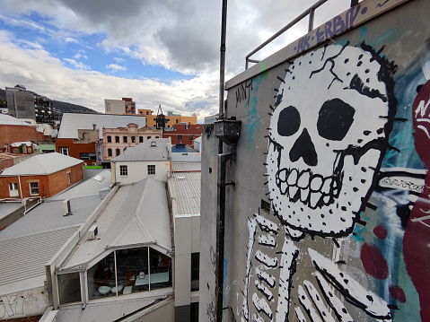 View of the city of Hobart from top of a building with a painted skeleton on the wall in Bidencopes Lane, Hobart’s street art hotspot. Hobart is the capital and most populous city of the island state of Tasmania, Australia.