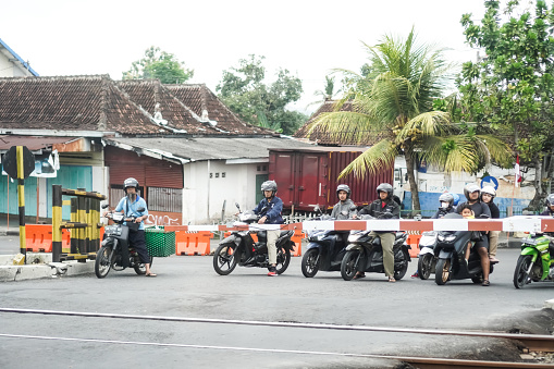 Java, Indonesia - August 19, 2023: People at Palang Pintu Kereta Api. Indonesian bikers and car drivers queued up behind railroad level crossing barrier and waiting the train passing during sunny day.