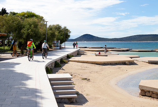 Vodice, Croatia - September 24, 2023: People walking and cycling at the promenade by the seaside beach in off-season sunset