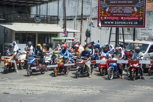 Java, Indonesia - August 19, 2023: People at Palang Pintu Kereta Api. Indonesian bikers and car drivers queued up behind railroad level crossing barrier and waiting the train passing during sunny day.