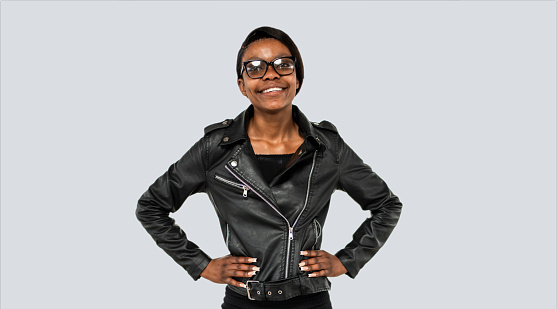 Portrait of a beautiful young african woman wearing black leather jacket and sunglasses standing with her hands on hips on grey background