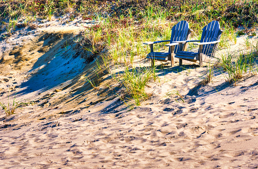 Two wooden Adirondack chairs rest on a grass covered dune on a Cape Cod beach on an early October afternoon.