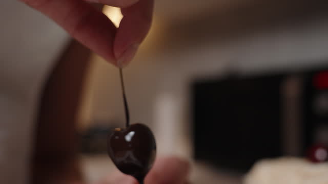 The woman covers the cherry on a stick with melted chocolate.