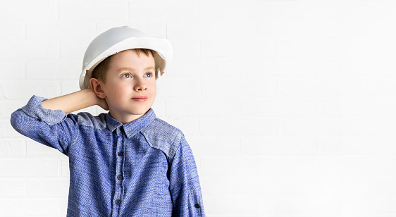 Child in a hardhat looks up and holds his hand behind head. Little boy engineer, architect, builder is thinking, looking for a new idea. Choice of profession, dreams concept. Copy space background.