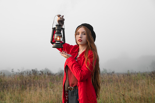 Woman with kerosene lamp in a red coat stands in a mystical misty morning field.