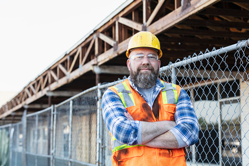 A mature man in his 50s with a beard, wearing a reflective vest, hardhat and safety goggles. The construction worker is standing with his arms crossed, looking at the camera. He is at a job site working on a strip mall renovation.