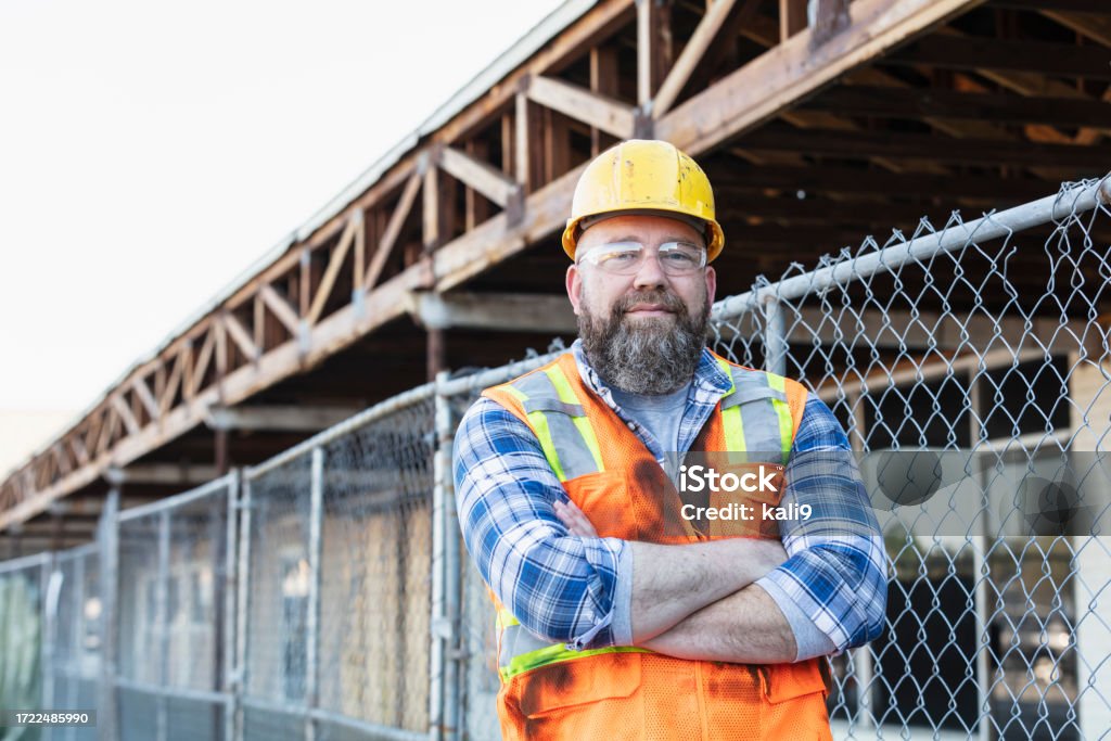 Construction worker next to building under renovation A mature man in his 50s with a beard, wearing a reflective vest, hardhat and safety goggles. The construction worker is standing with his arms crossed, looking at the camera. He is at a job site working on a strip mall renovation. Construction Worker Stock Photo