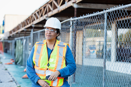 A mature Hispanic woman in her 40s wearing a reflective vest, hardhat and safety goggles. The construction worker is standing outdoors, holding a walkie-talkie, looking away from the camera. She is at a job site working on a strip mall renovation.