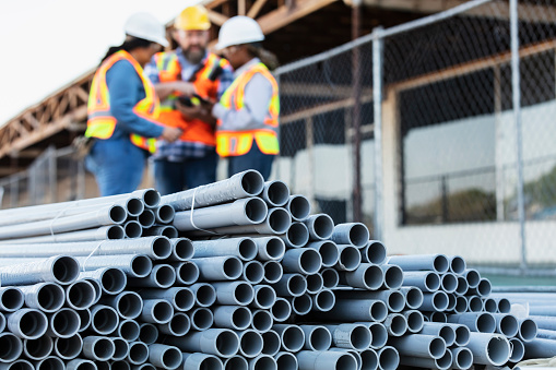 A stack of PVC pipes at a construction site, in the foreground. A group of construction workers are out of focus, having a meeting in the background. The project is the renovation of a strip mall.