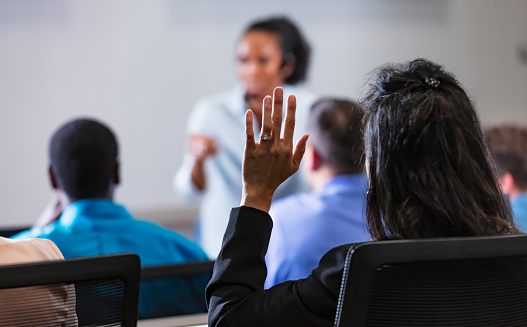 Over the shoulder view of a mature Asian woman in a class, seminar or business meeting, raising her hand to ask a question. The focus is on her hand in the foreground. The instructor and other members of the audience are out of focus in the background.