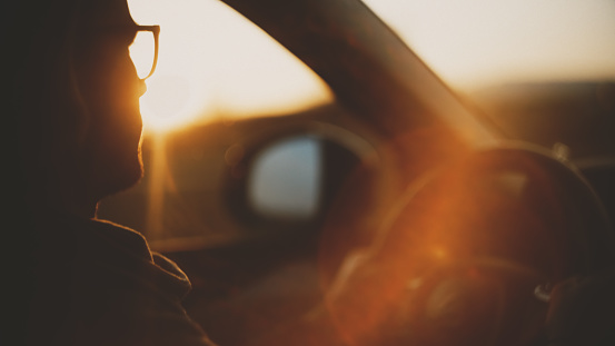 Silhouetted against the setting sun,a young man in eyeglasses drives with purpose,merging into the tranquil beauty of the twilight hour.