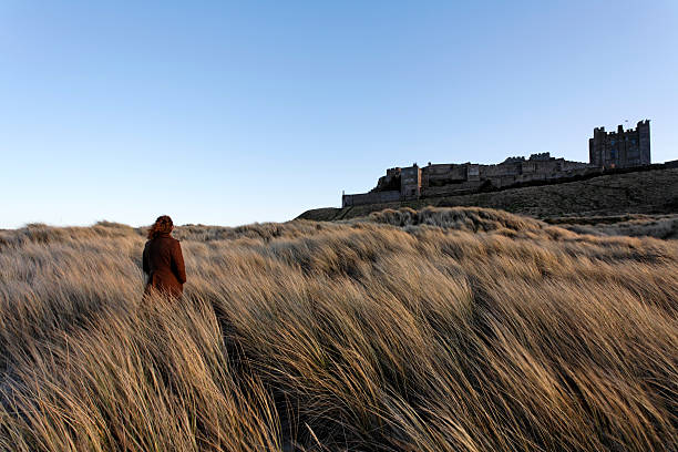 To the castle A lone woman walking towards a castle through grass covered dunes on a sunny evening Bamburgh stock pictures, royalty-free photos & images