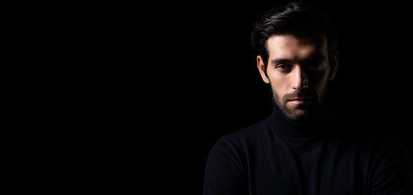 Smiling portrait of a young man looking at camera during a portrait session. Middle eastern man in casuals and curly hair sitting against black background.