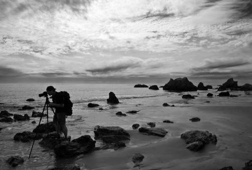 In black and white, a still photographer shoots stills at El Matador State Beach in Malibu, California against a dramatic sky.