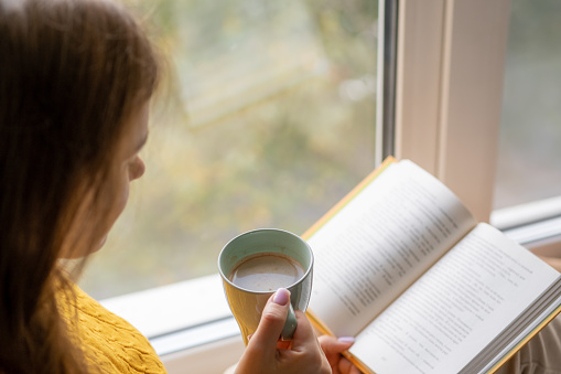 Young beautiful woman sitting by the window yellow knitted sweater read book, daily planner, notepad. Relax concept. Hold cappuccino glass of coffee with white foam. Text is out of focus