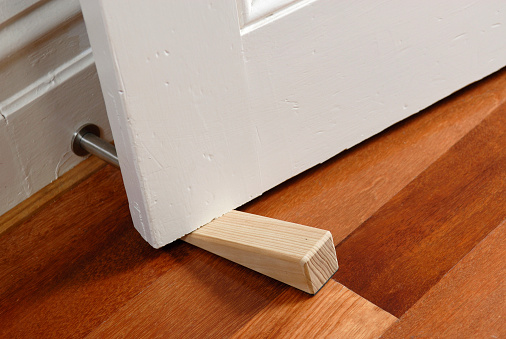 A wooden doorstop made of pine is being used to wedge open a door.  The door is an older style and the floor is timber / wood.
