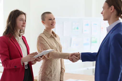 Woman with clipboard watches colleagues shaking hands in modern office. Happy employees make profitable business deal doing handshake