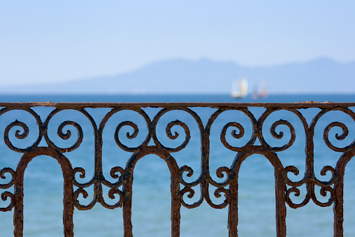 Ornate ironwork balcony fence railing positioned just below the horizon line featuring sailboats in an ocean bay, with low mountains and blue sky. The vista provides a beautiful scenic view of the horizon over water from Puerto Vallarta, Mexico. For concepts of tropical vacations and idyllic, relaxing tourist travel destinations. 