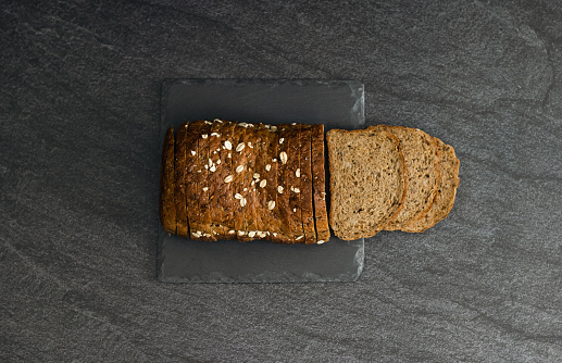 One dark rye bread with crushed oat grains and sunflower seeds lies cut into thin pieces lies on a stone stand in the center on a black background, flat lay close-up.The concept of baking bread.